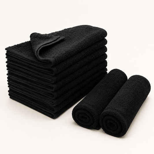 Ten black salon towels arranged as a folded stack and two rolls on a warm-white studio background