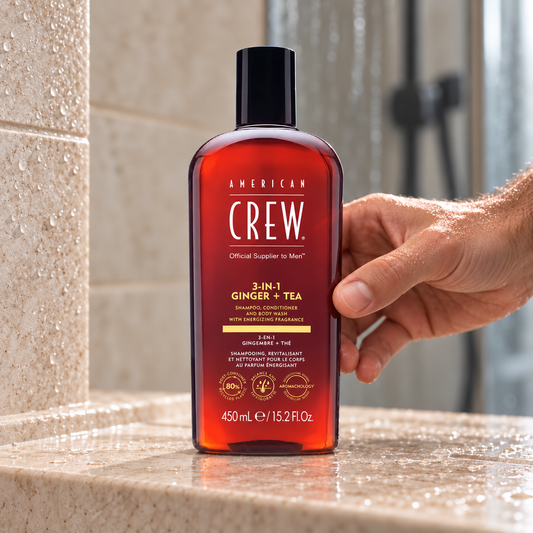 American Crew 3-in-1 Ginger + Tea 450ml bottle held on a wet shower ledge.