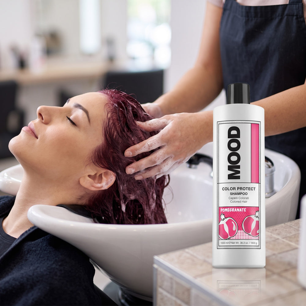 Stylist washing a client's red hair at a salon basin beside MOOD Color Protect Shampoo