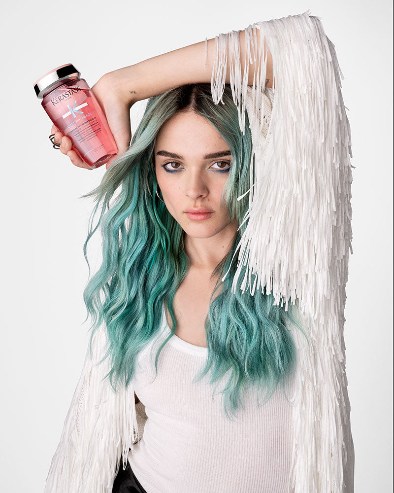 Woman with long teal-coloured hair holding a bottle of Kérastase Chroma Absolu shampoo