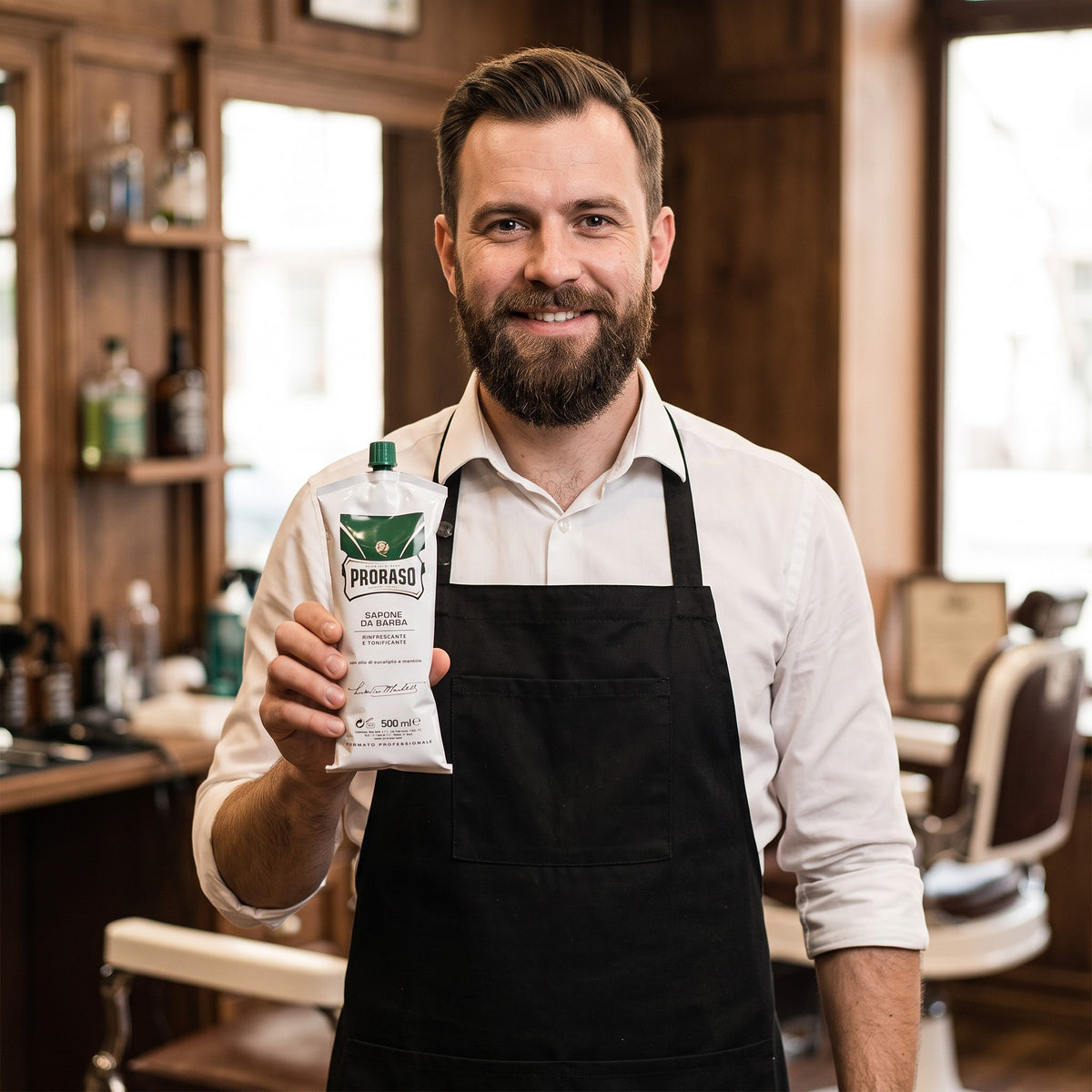 Smiling barber holding a Proraso Green Shaving Soap 500ml tube in a barbershop